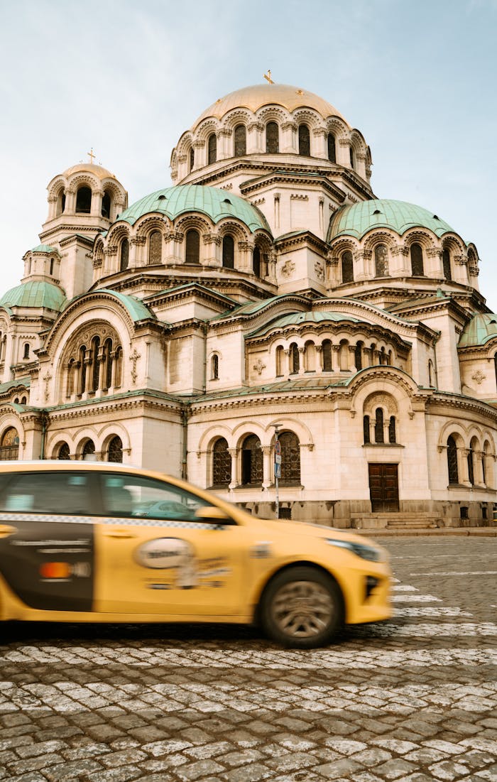 Yellow taxi passing by the iconic Alexander Nevsky Cathedral in Sofia, Bulgaria, under a clear sky.